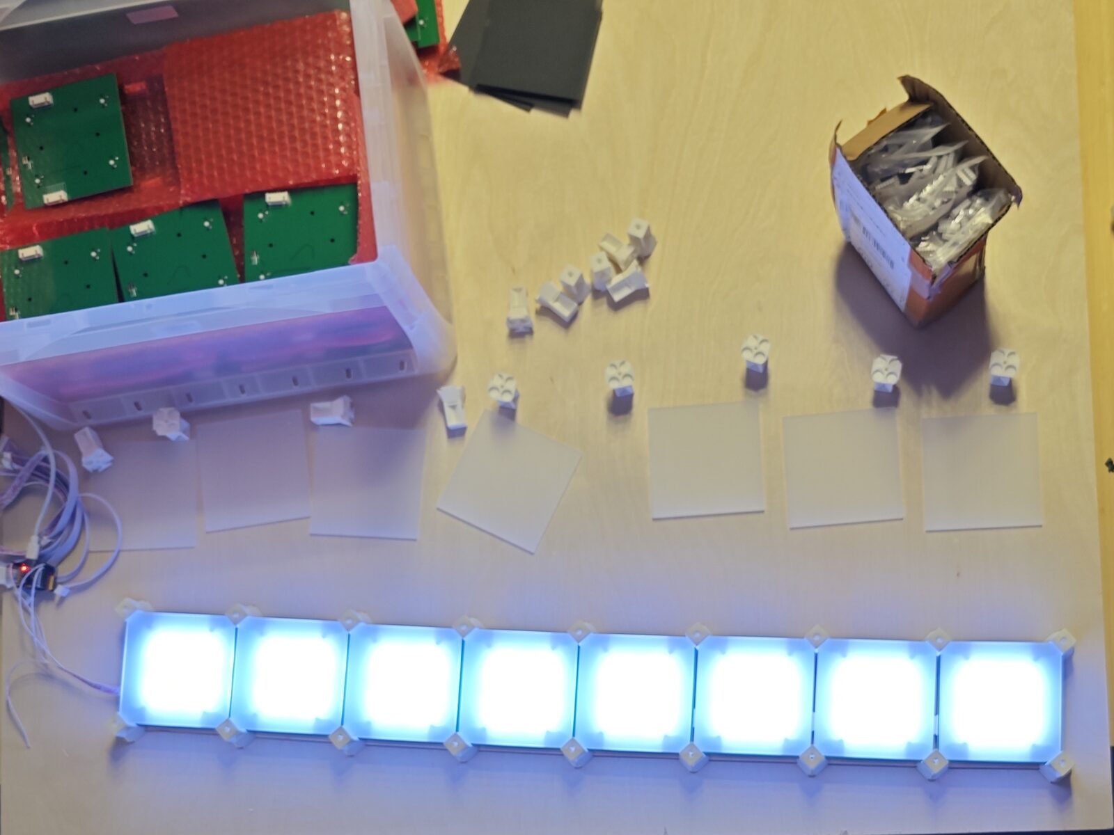 Bauteile der LED-Wand auf dem Tisch: grüne Platinen in einer Box, weiße Tastatur-Switches und Acrylplättchen, dazu eine Reihe aus acht hell blau-weiß leuchtenden LED-Modulen