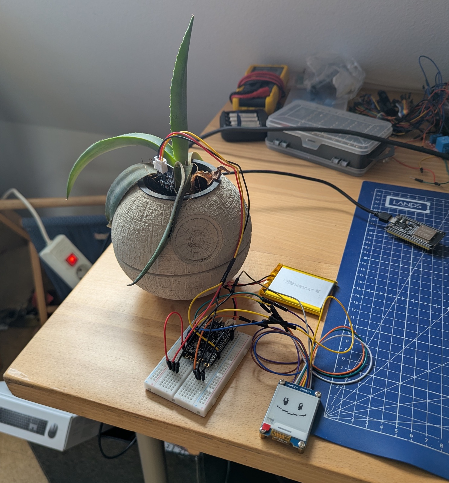 Aloe Vera in einem Todesstern-Blumentopf, daran ein ESP32 mit kapazitivem Sensor, ein LiPo-Akku, ein Breadboard und ein kleines E-Ink-Display mit lächelndem Gotchi-Gesicht