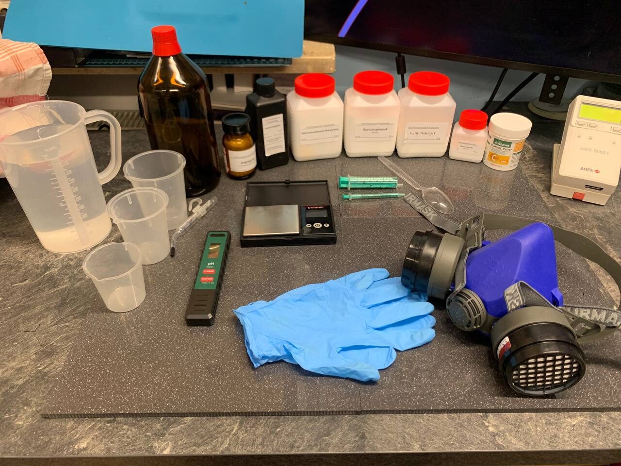 Selbst angemischte Foto-Chemie: Messbecher, braune und weiße Chemikalienflaschen, Feinwaage, pH-Meter, Spritzen, blaue Einweghandschuhe und eine Atemschutzmaske auf dem Labortisch