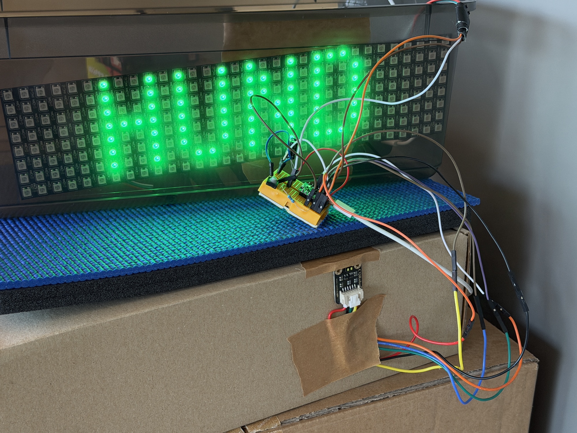 Die selbstgebaute Stoppuhr in Aktion: eine grüne 32×8-LED-Anzeige zeigt »WIAI«, darüber der Controller auf dem Steckbrett, vorn am Karton der Abstandssensor.