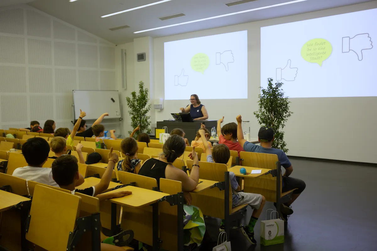 Kinder im Hörsaal strecken die Hände in die Höhe, vorne die Vortragende der Kinderuni.