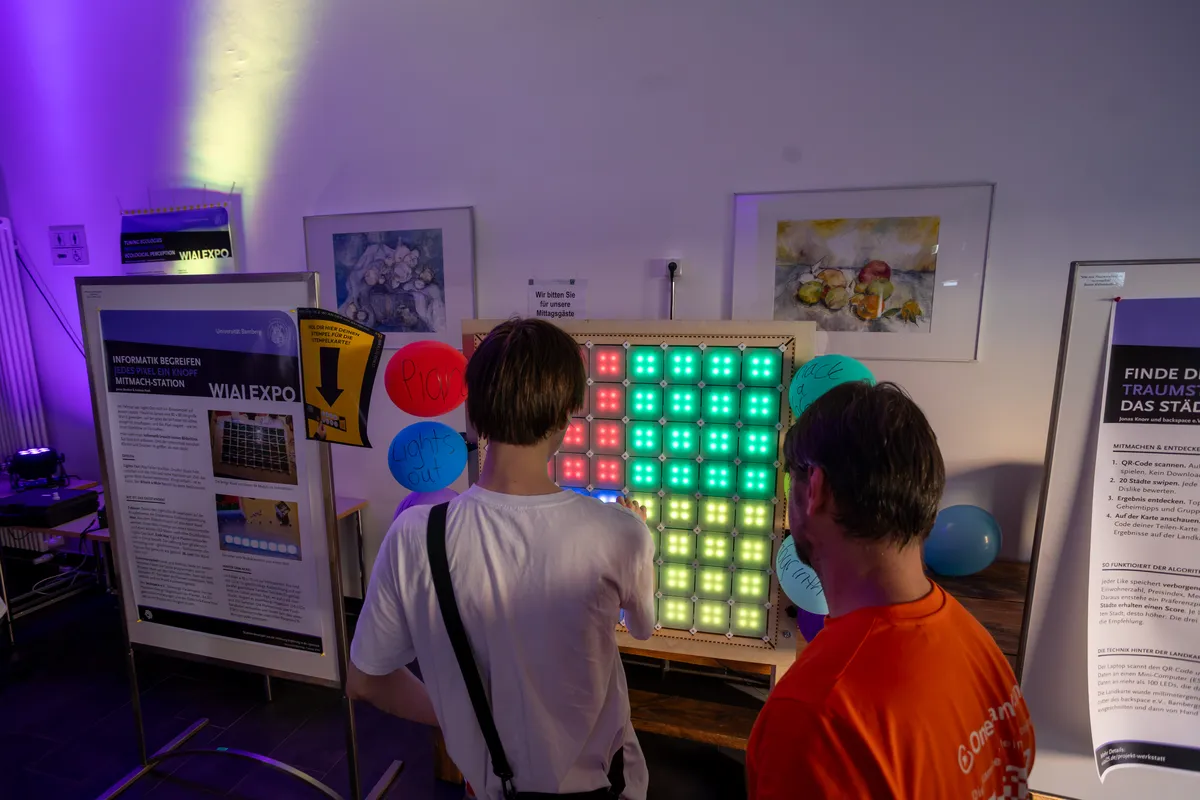 Zwei Gäste spielen an der bunt leuchtenden LED-Spiele-Wand, daneben das Stations-Poster und Luftballons