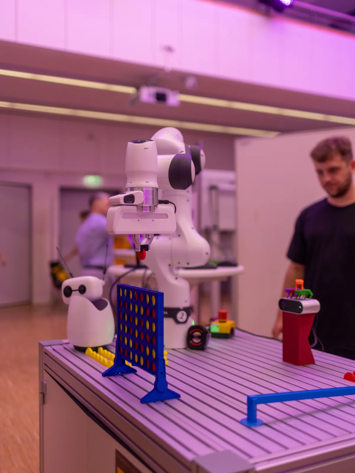 Ein weißer Roboterarm über einem blauen Vier-gewinnt-Brett, daneben wartet ein Gast.