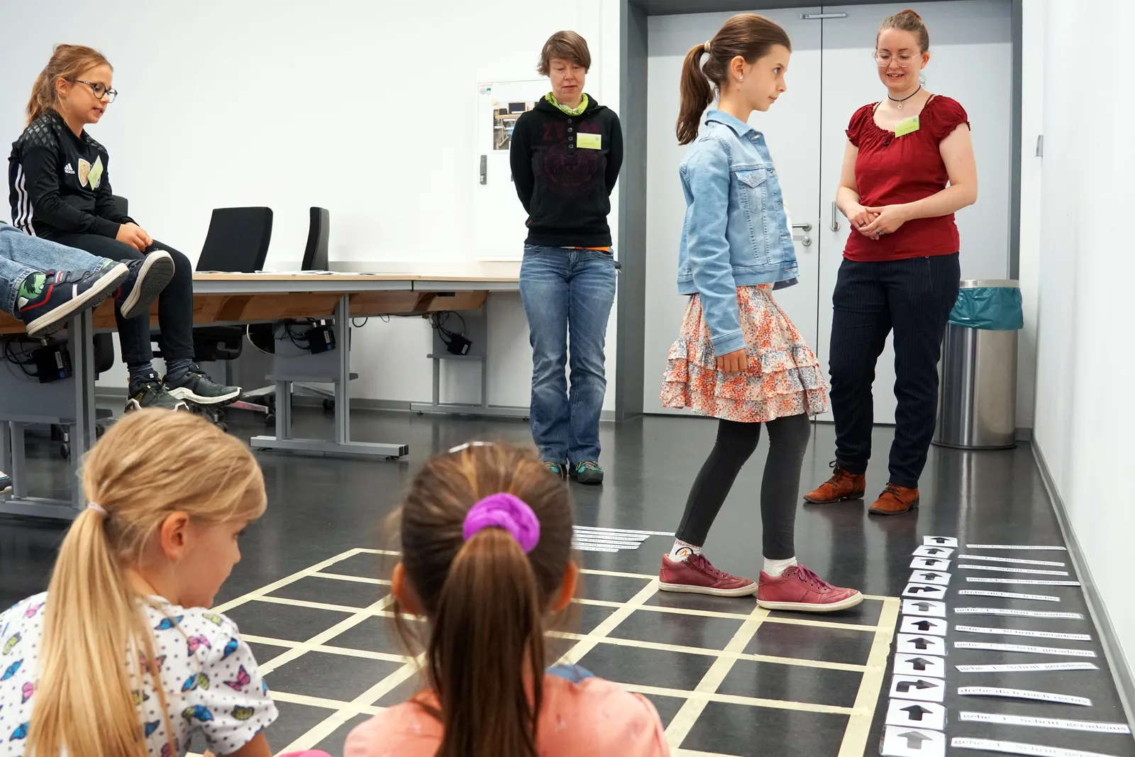 Ein Mädchen läuft beim Bamberger Informatiktag über ein auf den Boden geklebtes Raster, zwei Betreuerinnen und weitere Kinder schauen zu