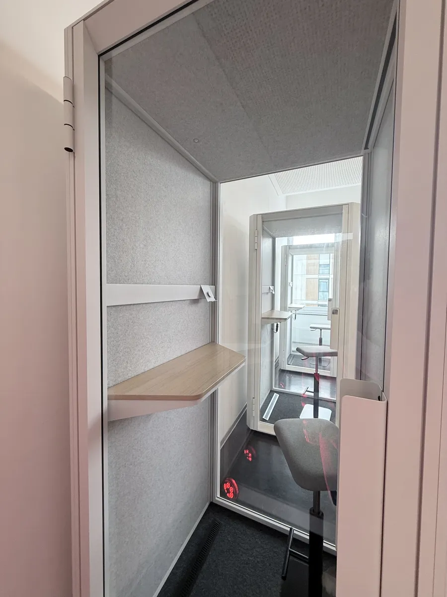 Blick durch die offene Glastür in eine schallisolierte Meeting-Box mit grauen Akustikwänden, Holzablage und Stuhl