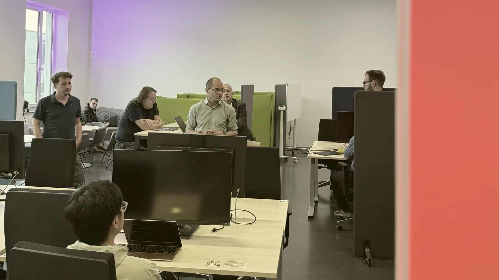 Blick in den neuen Study Space der WIAI: Studierende und Mitarbeiter an Arbeitsplätzen mit Monitoren, grüne Akustikmöbel, rechts die rote Wand einer Meeting-Box